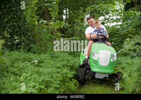 Père et fils : un père laisse son fils de 5 ans à diriger le tracteur-tondeuse qu'il coupe l'herbe Banque D'Images