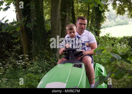 Père et fils : un père laisse son fils de 5 ans à diriger le tracteur-tondeuse qu'il coupe l'herbe Banque D'Images