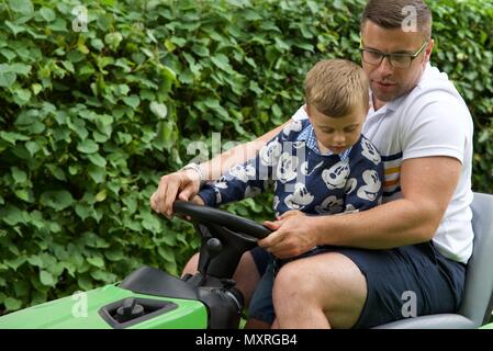 Père et fils : un père laisse son fils de 5 ans à diriger le tracteur-tondeuse qu'il coupe l'herbe Banque D'Images