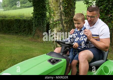 Père et fils : un père laisse son fils de 5 ans à diriger le tracteur-tondeuse qu'il coupe l'herbe Banque D'Images