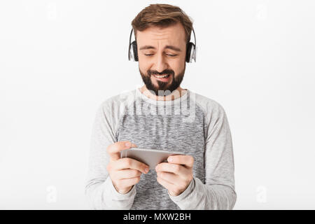 Photo de jeune homme excité nerveux isolated over white wall background jouer des jeux par téléphone portable à côté portant des écouteurs. Banque D'Images