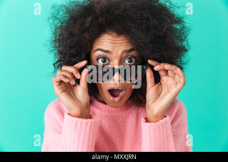 Photo de femme surprise 20s avec des cheveux hirsutes à la recherche sur l'appareil photo avec l'excitation du dessous des lunettes noires sur fond bleu isolé Banque D'Images