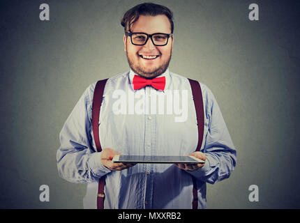 L'homme corpulent élégant noeud papillon en montrant tablet heureusement smiling at camera sur fond gris Banque D'Images