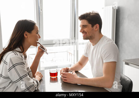 Image homme et femme à l'un l'autre tout en restant assis à table à l'intérieur près grande fenêtre et manger du chocolat avec du thé Banque D'Images