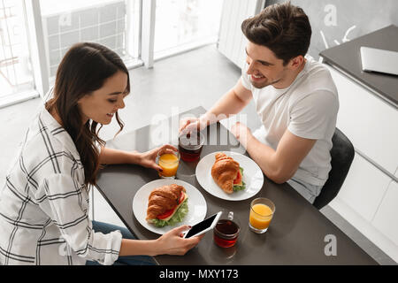 Image homme et femme assis à table à l'intérieur près grande fenêtre et prendre le petit-déjeuner lors de l'utilisation de téléphone mobile Banque D'Images