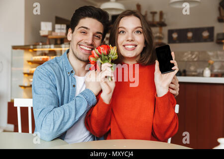 Photo de smiling couple sitting at table in cafe confortable tout en woman holding de belles fleurs et smartphone en mains Banque D'Images