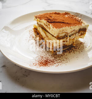 Gros plan d'une tranche de tiramisu traditionnel fait maison (dessert italien) sur une plaque blanche avec une cuillère sur un en conseil. Format carré. Banque D'Images