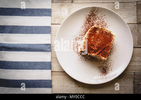 Vue de dessus d'une coupe de tiramisu traditionnel fait maison (dessert italien) sur une assiette blanche sur une planche en bois avec un chiffon de cuisine blanc et bleu. Banque D'Images