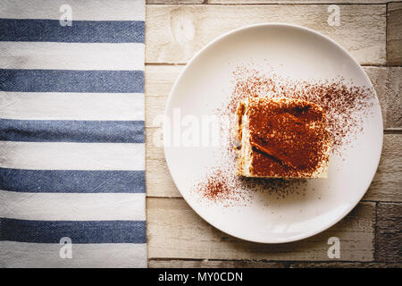 Vue de dessus d'une coupe de tiramisu traditionnel fait maison (dessert italien) sur une assiette blanche sur une planche en bois avec un chiffon de cuisine blanc et bleu. Banque D'Images