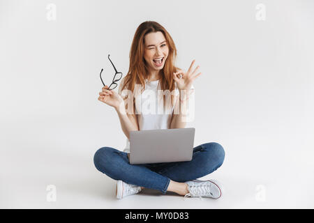 Femme joyeuse dans t-shirt assis sur le plancher avec un ordinateur portable tout en montrant ok s'identifier et avec des clins d'oeil au-dessus de l'appareil photo arrière-plan gris Banque D'Images