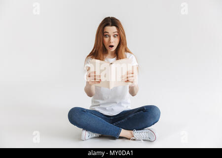 Femme surprise en t-shirt assis sur le plancher et de la lecture livre avec la bouche ouverte sur fond gris Banque D'Images