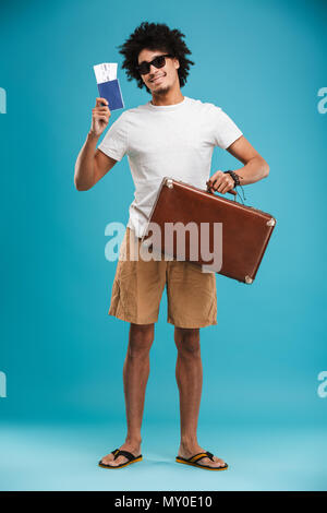 Photo de young man bouclés de l'Afrique de l'isolated over blue wall background munis de billets avec passeport et valise. Banque D'Images