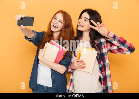 Portrait de deux jolies jeunes filles adolescentes de l'école tenue des cahiers et en tenant un isolé sur fond jaune selfies Banque D'Images