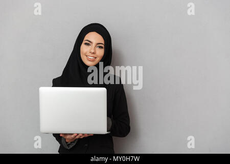 Portrait of smiling woman arabe 20s en noir hijab traditionnel looking at camera while holding ordinateur portable isolé sur fond gris Banque D'Images