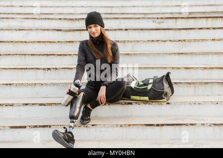 Photo de femme handicapée smiling in sportswear avec prothèse assis à l'escalier d'extérieur et de la tenue de la coupe du thermos Banque D'Images