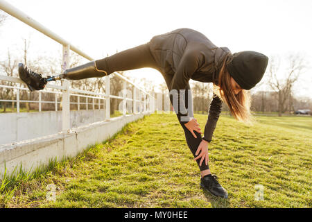 Photo de mobilité tournant femme dans Vêtement de ski et faire des étirements prothèse sur l'herbe à l'aide de garde-fous Banque D'Images