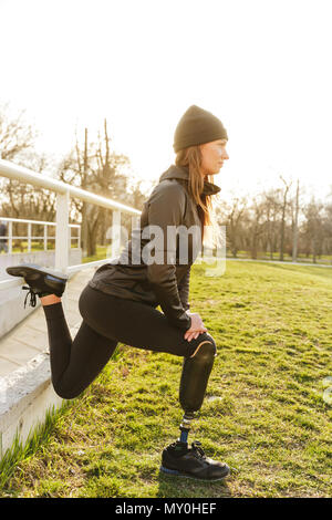 Photo de mobilité en cours d'exécution dans les vêtements de sport femme accroupie et stretching prothèse sur l'herbe à l'aide de garde-fous Banque D'Images