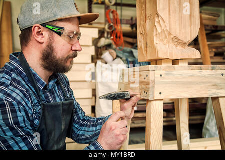 Menuisier expérimenté dans les vêtements de travail et de petits buiness propriétaire travaillant dans l'atelier de menuiserie, à l'aide d'un marteau frappant les ongles, sur la table est un marteau et Banque D'Images