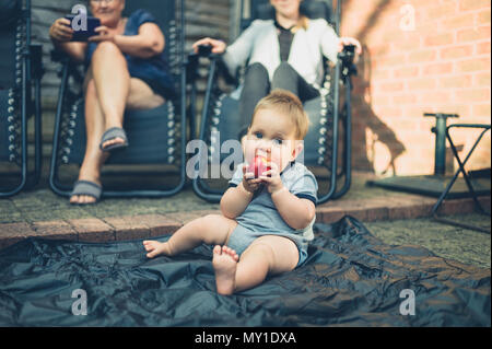 Un mignon petit bébé est assis sur le sol dans le jardin et est en train de manger une pêche Banque D'Images