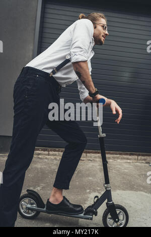 Vue latérale d'un jeune homme portant des lunettes et des bretelles riding scooter Banque D'Images