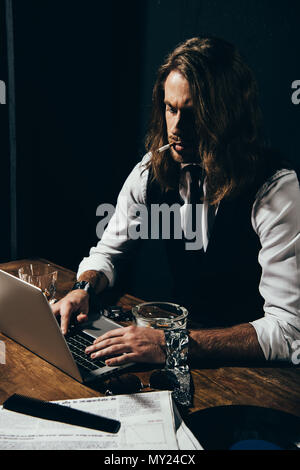 Jeune homme aux cheveux longs en tenue de cérémonie cigarette et typing on laptop Banque D'Images