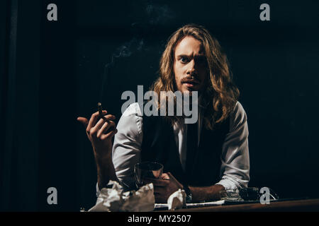 Beau barbu homme aux cheveux longs en tenue de cérémonie de fumer un cigare tout en maintenant le verre de whisky et looking at camera Banque D'Images