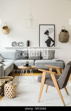 Fauteuil gris à côté de la table en bois en intérieur lumineux avec poster et coussins sur un canapé. Photo réelle Banque D'Images