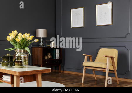 Armoire en bois dans le coin d'une salle de séjour avec boiseries d'intérieur avec des affiches sur le mur à côté d'un fauteuil et table jaune avec des tulipes. Banque D'Images