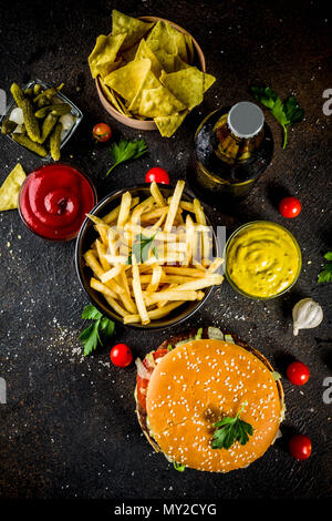 Divers cuisine de fête, hamburgers, frites, croustilles de pommes de terre, concombres, oignons, tomates et les bouteilles de bière froide, fond en béton noir rouillé Banque D'Images