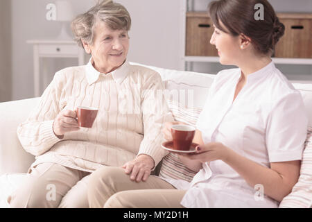 Femme âgée et son soignant privé assis ensemble sur un canapé, boire le thé à la maison Banque D'Images
