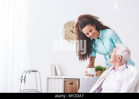 Le fournisseur de jeunes servant une tasse de thé à un heureux, homme plus âgé dans une maison de retraite Banque D'Images