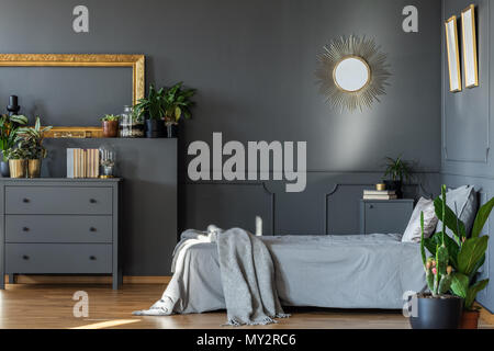 Miroir décoratif accroché au mur gris foncé de l'intérieur chambre à coucher avec des plantes fraîches et des livres sur l'armoire en bois Banque D'Images