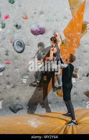 Long Shot of young woman escalade un mur avec prises avec l'homme soutenant par derrière Banque D'Images