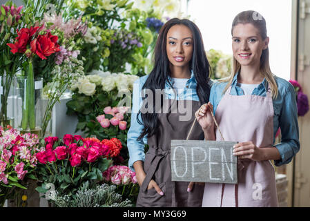 Beautiful smiling jeunes fleuristes multiethnique holding open sign and smiling at camera in flower shop Banque D'Images