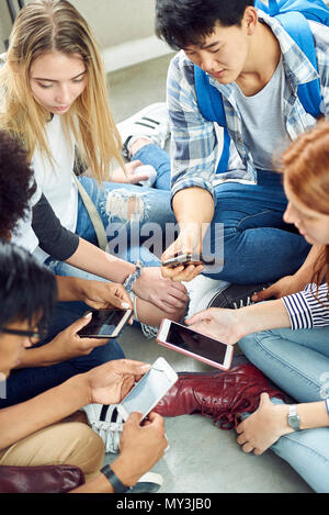 Groupe d'étudiants assis ensemble à l'étage, à la recherche de téléphones intelligents Banque D'Images