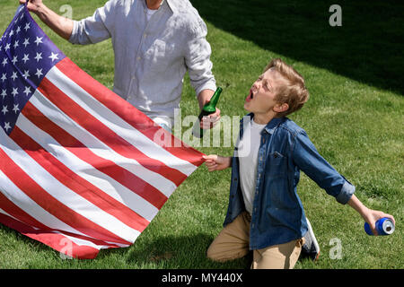 Père et fils assis sur l'herbe avec nous drapeau, garçon crier et holding de soude peut, le jour de l'indépendance de l'Amérique concept Banque D'Images