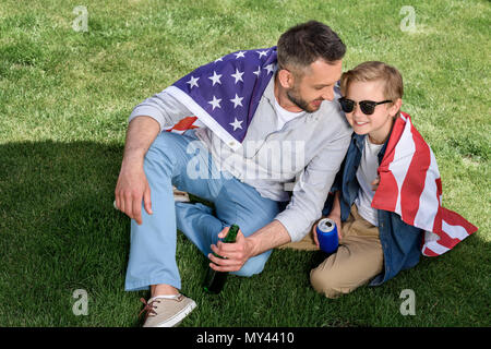 Père et fils assis sur l'herbe avec nous d'un drapeau et d'organiser la bière et soda, jour de l'indépendance de l'Amérique du concept Banque D'Images