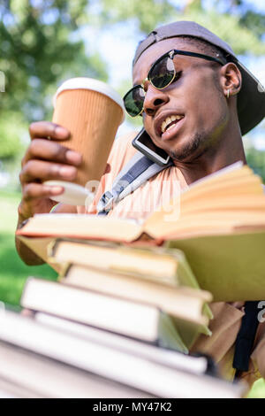 Young african american man talking on smartphone pendant que holding pile of books et tasse à café jetable Banque D'Images