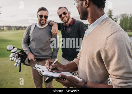 African American man écrit dans le presse-papiers tout en regardant sur deux joueurs de golf Banque D'Images