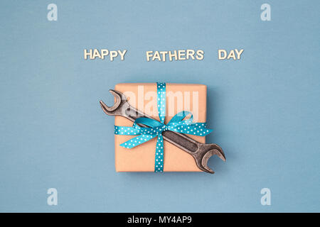 Happy Father's Day card avec boîte-cadeau enveloppé dans du papier kraft et attaché avec du ruban bleu à pois blancs et clé sur fond gris bleu. Carte postale avec des lettres en bois. Banque D'Images