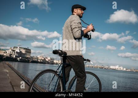 L'homme élégant avec appareil photo et film vintage bike debout sur la rive de la rivière Banque D'Images