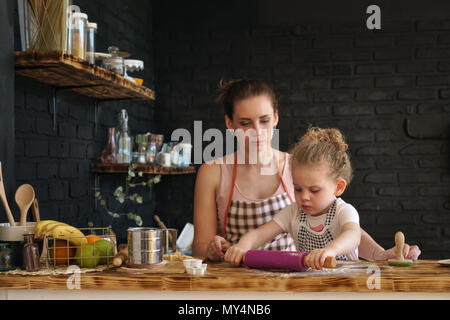 Jeune mère et fille préparer des cookies dans la cuisine. Ils sont dans des tabliers. Petite fille roule la pâte avec un rouleau à pâtisserie. Little Helper. Le temps passé en famille. Banque D'Images