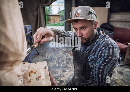 Jeune homme dans un menuisier scies vêtements de travail à la tête d'un homme avec un arbre , à l'aide d'un burin dans l'atelier, autour d'un lot d'outils pour le travail Banque D'Images