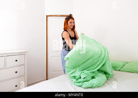 Сlose de jeunes femme en jeans et t-shirt noir nettoie le lit de vert-blanc literie géométrique dans une chambre avec des murs blancs et d'un coffre à dr Banque D'Images