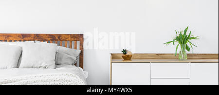 Close-up d'une partie d'un lit avec draps et oreillers et une armoire avec les fleurs coupées dans un vase sur le haut standing contre un mur blanc dans un Banque D'Images