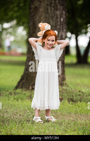 Jolie petite fille aux cheveux rouge en robe blanche à teddy bear park Banque D'Images