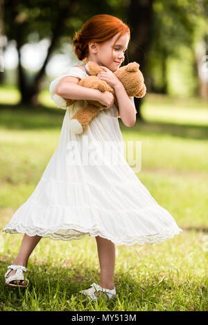 Heureux redhead girl in white dress hugging teddy bear at park Banque D'Images