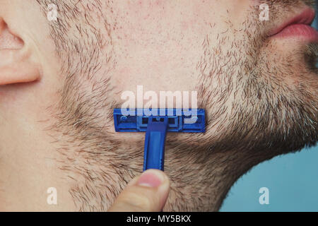 Vue d'un jeune homme barbu avec un rasoir brutal. visage sur le fond bleu. grandes soies rugueuses sur la peau velue. Vue de côté close up Banque D'Images