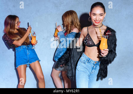 La mode smiling asian woman holding glass avec cocktail tandis que les amis de boire des cocktails et à l'aide derrière smartphone at party Banque D'Images