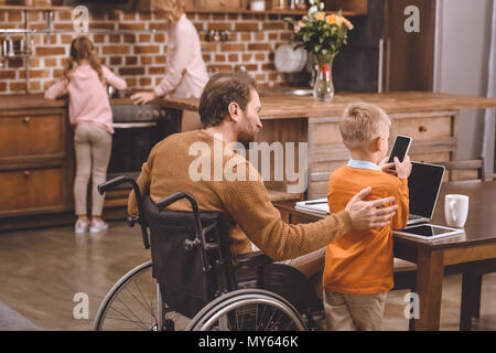 Vue arrière de père en fauteuil roulant et à mobilité réduite petit-fils à l'aide d'un ordinateur portable et smartphone ensemble à la maison Banque D'Images